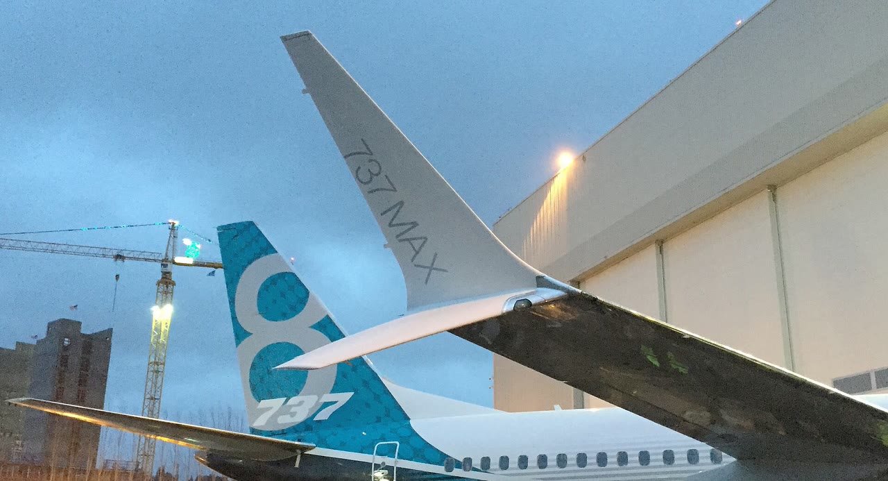 Ponta de asa de um Boeing 737 MAX com a inscrição 737 MAX e, atrás dela, a cauda azul e branca do avião, diante de um hangar ao entardecer