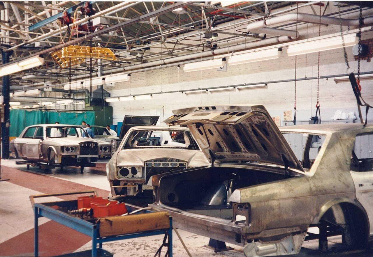 Carrocerias de sedãs ainda sem pintura, de metal exposto, em preparação numa linha da fábrica de Crewe, com trilhos e lâmpadas no teto